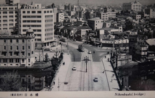 【32】国内諸街道の元標。日本橋は、中央区日本橋一丁目と室町一丁目との間にある日本橋川に架かるコンクリート造りの橋である。1603年（慶長8年）江戸幕府が開かれたとき日本橋、京橋一帯の新市街ができ、日本橋はそのとき初めて架けられたもので、翌年には日本橋を起点として諸街道の里程を定め、一里毎に一里塚が置かれた。1873年（明治6年）に「東京は日本橋。京都は三条の中央を以て国内諸街道の元標とす」と定められ、今も橋の中央に道路元標の鉄塔が建っている。現在の橋は1911年（明治44年）架け替えられたものである。1964年（昭和39年）東京オリンピックの前年、日本橋川を塞ぐように首都高速道路が建設され本来の景色を失ってしまった。戦災復興による道路整備が計画どおりに進まず、五輪へ向けて都心部の交通混雑を回避するため川や沿岸部などの空間を利用しての整備だった。その後、日本の高度経済成長を支える重要インフラとして活躍したが、すでに50年が経過して更新時期を迎え、景観を取り戻す署名運動も行われ、地下化が検討されている。【32】1955年（昭和30年）頃・（東京名所）日本橋