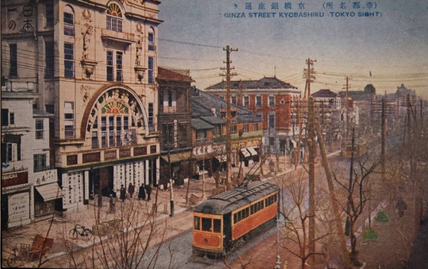 【5】資生堂×辰野金吾。文化演出、画廊の開花。大正時代の銀座通り。官主導、ウォートスによるジョージアン様式の赤煉瓦街の均質な街並みに、塔や時計が取り付けられる変化が起きた。高い文化意識を備えた銀座人が台頭し、現代に通じる気品と格式ある都市空間が創造された。単に銀座で商品を売ることにとどまらず、文化人として建てる建築にこだわりを見せた。その代表格が、調剤薬局からスタートした資生堂である。初代社長であり写真家でだった福原信三は、1916年（大正5年）に、東京駅を設計し明治の建築界に君臨した近代建築の父、辰野金吾と交流を持ち、煉瓦造3階建の化粧品部ビルを完成させる。絵葉書の中央に描かれている丸窓のある建築で、1925年（大正14年）パリ万博で発表される以前に、アール・デコを予感させる装飾を持ち込んだ。3年後には資生堂ギャラリーを開設し、芸術家を発掘紹介する文化事業もスタートさせる。その後、日動画廊をはじめ多くの画廊が街中に開廊し、芸術鑑賞、文化演出の場として銀座を開花させたのである。【5】1921年（大正10年）頃・（帝都名所）京橋銀座通り