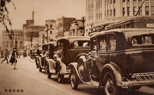 【19】新聞、報道の街。雑誌『主婦之友』に附録として発行された72枚のうちの１枚。以下解説文より「銀座通りといふのは、北は京橋の袂から、南は新橋驛附近までの総称で、そのその中心をなすのが、銀座四丁目、即ち尾張町の交叉點であります。右手の白い建物は、新装成つた服部時計店で、左手に見えるのが、東京朝日新聞社です。」。明治時代、煉瓦街の建設に伴い、東京日日新聞、東京朝日新聞、読売新聞や國民新聞など新聞社のほとんどが銀座に集中し、印刷業や広告代理店なども集中した時期があった。しかし関東大震災により壊滅的打撃を受けると、読売が大手町へ移転するなど銀座への集中も途絶えることとなった。また、日本電報通信社（電通）が銀座に設立され、広告原稿の受け渡しの利便性から地方新聞社の多くが銀座周辺に東京支局を構えた。そのため現在でも支局や支社を構えるところが存在する。【19】1932年（昭和7年）・銀座尾張町