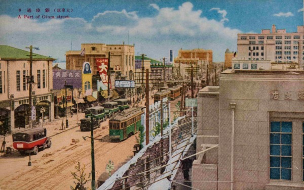 【27】街を往く人たちが、車窓の景色だった都電。1882年（明治15年）馬車鉄道が新橋〜日本橋間で開業、1903年東京電車鉄道と名を変え電化し、数寄屋橋〜神田橋間に東京市街鉄道が開業した。後に東京市電として公営化し、路線・系統の拡大がされた。絵葉書に描かれているのは昭和10年頃に登場したダーク・グリーンの車体。真鍮の金具を光らせていた。アール・ヌーボーの意匠が施された街路灯、 商店のショーウインドウにも電気のイルミネーションが使われ話題を集めた。1934年（昭和9年）、東京地下鉄道によって敷設された地下鉄（現在の東京メトロ銀座線）が銀座まで延伸し、1950年代より整備が急速に進み、西銀座駅の丸ノ内線が開業。その一方で自動車の増加に伴い、惜しまれつつも都電は昭和42年に廃止された。【27】1940年（昭和15年）頃・（大東京）銀座通り