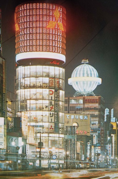 【37】高度経済成長期に始まった歩行者天国。1964年（昭和39年）東京オリンピックが開催され、1969年（昭和44年）GNP世界第2位となるなど、高度経済成長の時代に。1965年（昭和38年）完成した三愛ドリームセンタービル。リコー創業者市村清が戦後間もない1946年（昭和21年）旧六十九銀行跡地に2階建のビルを建設し、以後この地から経営拡大。法隆寺五重塔をヒントに「建物中心に大きな柱を立ててビル全体を総ガラス化させた円筒型のビル」を考案、名称も一般公募で決定した。外壁には社章である眼鏡のマークが配してあった。三菱電機スカイリングのネオンも長年親しまれた。その他にも、銀座最大を誇った東芝ビル、銀座ライオンビル、名鉄メルサ、資生堂ザ・ギンザなど、新しいビルが次々と建てられた。中央通りでは、1970年（昭和45年）歩行者天国が実施されるようになり休日の定番となる。【37】1965年（昭和38年）頃・三愛ビルネオン