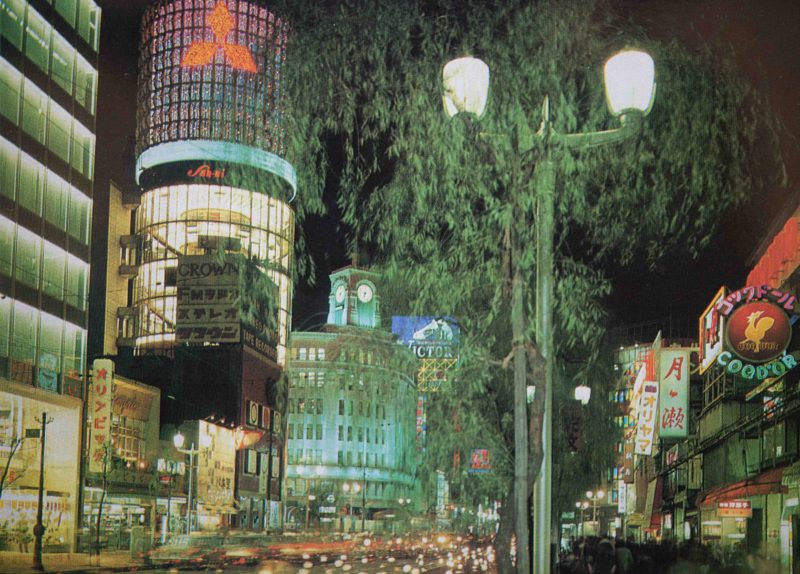 【38】高度経済成長期に始まった歩行者天国。1964年（昭和39年）東京オリンピックが開催され、1969年（昭和44年）GNP世界第2位となるなど、高度経済成長の時代に。1965年（昭和38年）完成した三愛ドリームセンタービル。リコー創業者市村清が戦後間もない1946年（昭和21年）旧六十九銀行跡地に2階建のビルを建設し、以後この地から経営拡大。法隆寺五重塔をヒントに「建物中心に大きな柱を立ててビル全体を総ガラス化させた円筒型のビル」を考案、名称も一般公募で決定した。外壁には社章である眼鏡のマークが配してあった。三菱電機スカイリングのネオンも長年親しまれた。その他にも、銀座最大を誇った東芝ビル、銀座ライオンビル、名鉄メルサ、資生堂ザ・ギンザなど、新しいビルが次々と建てられた。中央通りでは、1970年（昭和45年）歩行者天国が実施されるようになり休日の定番となる。【38】1970年頃・三愛ビルネオン、銀座和光と柳